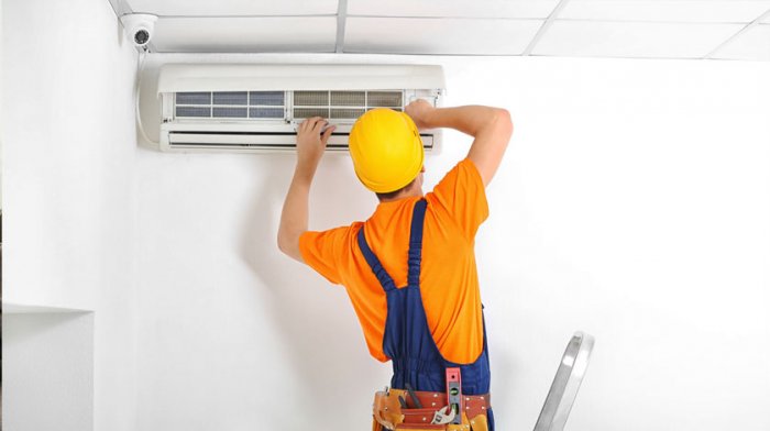 A technician wearing a yellow hard hat and orange shirt is installing or repairing an air conditioning unit on a white wall, ensuring optimal performance by Season Control.