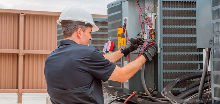 A technician wearing a hard hat and gloves is working on an HVAC unit, using electrical tools and meters to ensure optimal by season control of the equipment.